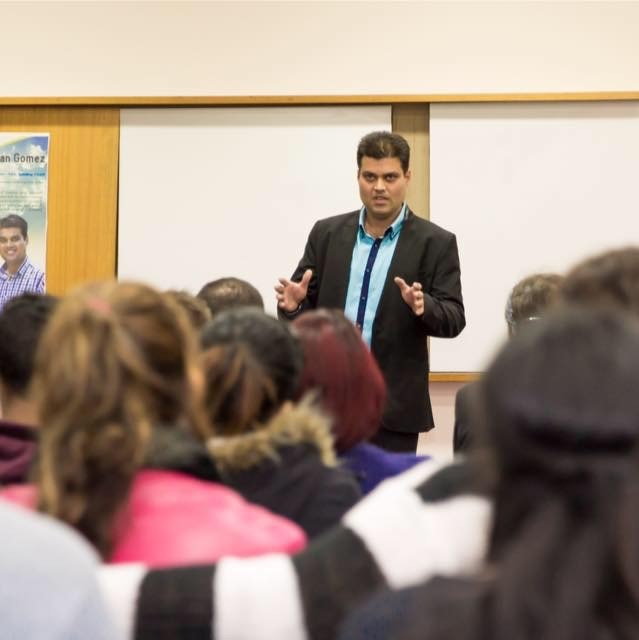 Ryan Gomez speaking passionately to a packed lecture hall in black blazer — gesturing while addressing a diverse audience