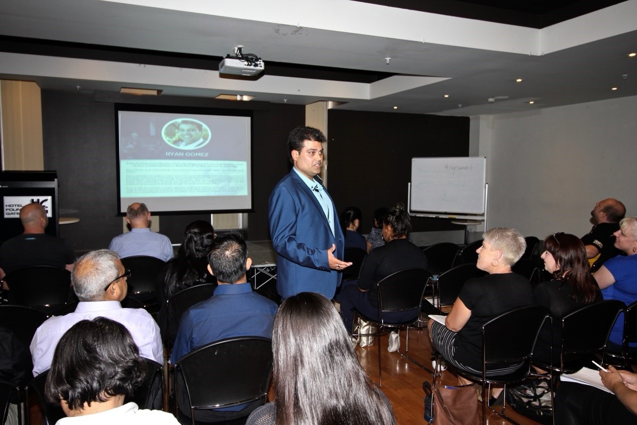 Ryan Gomez presenting with his banner at Hotel Fountain Gate — blue suit, engaging a full room during a leadership seminar